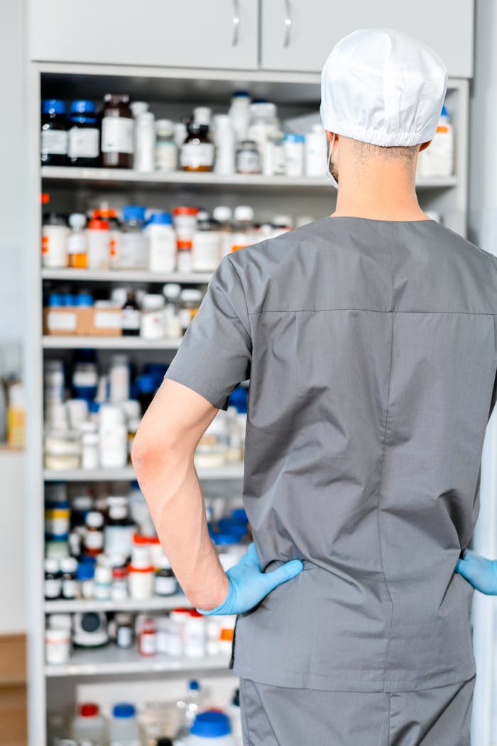ours-journey Male pharmacist in surgical cap and gloves organizing shelves of medication in a pharmacy.